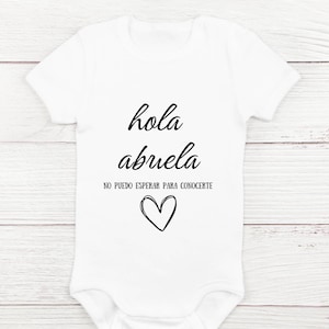 May include: White baby onesie with black text that reads "hola abuela" and "no puedo esperar para conocerte" with a black heart below the text.