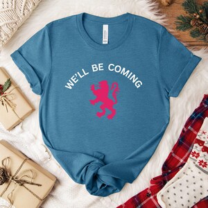 May include: A teal crew neck t-shirt with the text "WE'LL BE COMING" in white above a pink lion graphic. The shirt is folded, and the image includes wrapped gifts and plaid pajamas.