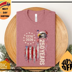 Pode incluir: Camiseta rosa empoeirado com um design de bandeira americana desgastada, uma cabe&ccedil;a de &aacute;guia e o texto "1776 2026 250 YEARS". A camiseta tem gola redonda e &eacute; feita de material macio.