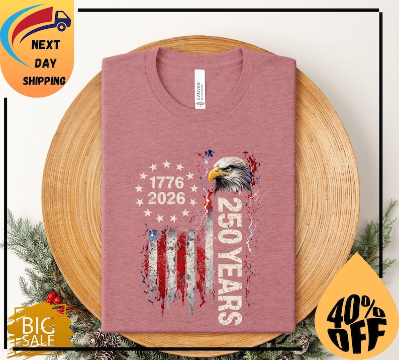 Pode incluir: Camiseta rosa empoeirado com um design de bandeira americana desgastada, uma cabe&ccedil;a de &aacute;guia e o texto "1776 2026 250 YEARS". A camiseta tem gola redonda e &eacute; feita de material macio.