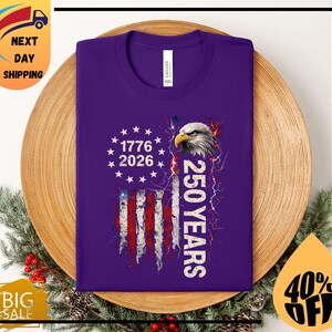 Pode incluir: Uma camiseta roxa com um design da bandeira americana, o texto "1776 2026" e "250 YEARS". O design inclui uma cabe&ccedil;a de &aacute;guia e raios. A camiseta &eacute; exibida em um prato de madeira com decora&ccedil;&otilde;es festivas.