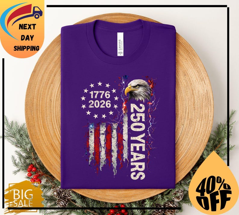Pode incluir: Uma camiseta roxa com um design da bandeira americana, o texto "1776 2026" e "250 YEARS". O design inclui uma cabe&ccedil;a de &aacute;guia e raios. A camiseta &eacute; exibida em um prato de madeira com decora&ccedil;&otilde;es festivas.