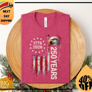 Pode incluir: Uma t-shirt rosa com um design gr&aacute;fico da bandeira americana, uma cabe&ccedil;a de &aacute;guia, os anos 1776 e 2026 e o texto "250 YEARS". A t-shirt &eacute; apresentada numa superf&iacute;cie de madeira.