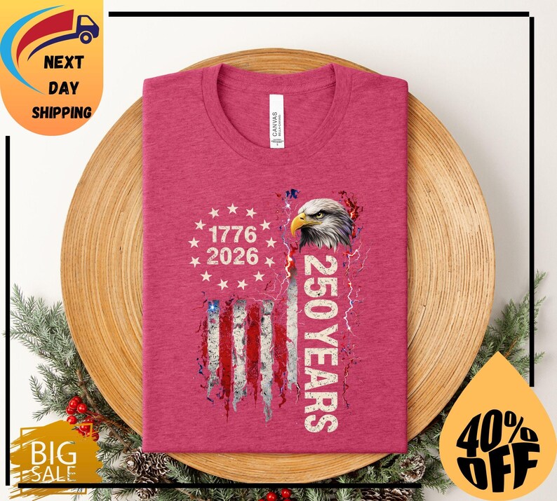 Pode incluir: Uma t-shirt rosa com um design gr&aacute;fico da bandeira americana, uma cabe&ccedil;a de &aacute;guia, os anos 1776 e 2026 e o texto "250 YEARS". A t-shirt &eacute; apresentada numa superf&iacute;cie de madeira.