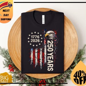 Pode incluir: Camiseta preta com um design da bandeira americana, o texto "1776 2026" e "250 YEARS". O design inclui uma cabe&ccedil;a de &aacute;guia e raios. A camiseta &eacute; exibida em um prato de madeira com decora&ccedil;&otilde;es festivas.