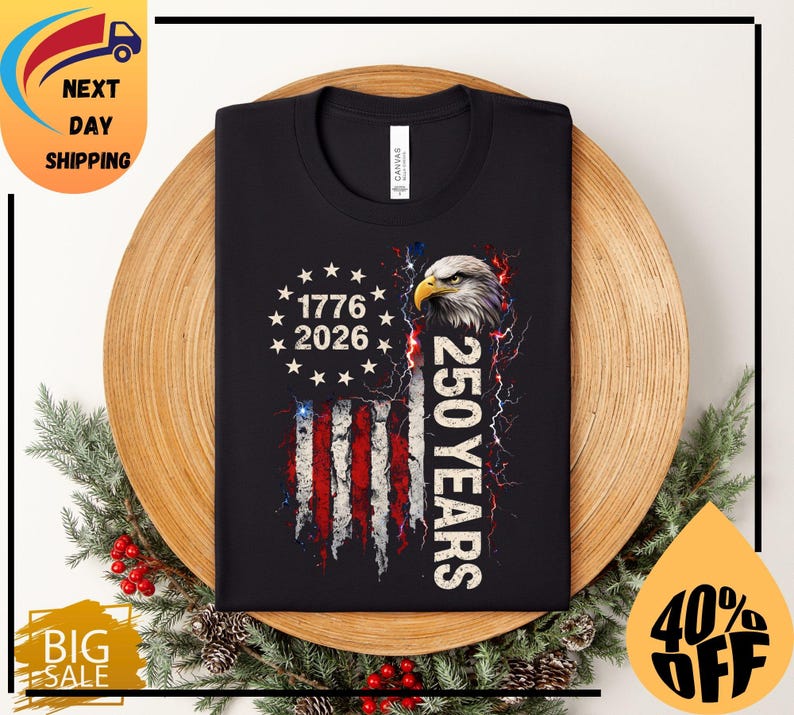 Pode incluir: Camiseta preta com um design da bandeira americana, o texto "1776 2026" e "250 YEARS". O design inclui uma cabe&ccedil;a de &aacute;guia e raios. A camiseta &eacute; exibida em um prato de madeira com decora&ccedil;&otilde;es festivas.