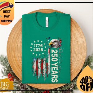 Pode incluir: Uma t-shirt verde com um design patri&oacute;tico. O design inclui uma bandeira americana, o texto "1776 2026" e "250 YEARS", juntamente com uma ilustra&ccedil;&atilde;o de uma &aacute;guia. A t-shirt &eacute; apresentada num prato de madeira.