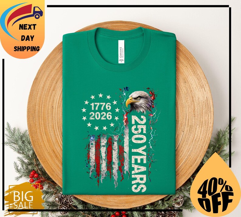 Pode incluir: Uma t-shirt verde com um design patri&oacute;tico. O design inclui uma bandeira americana, o texto "1776 2026" e "250 YEARS", juntamente com uma ilustra&ccedil;&atilde;o de uma &aacute;guia. A t-shirt &eacute; apresentada num prato de madeira.