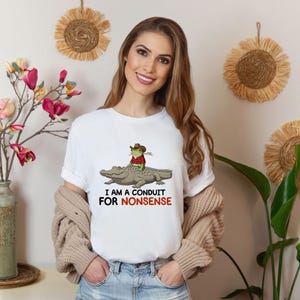 May include: White t-shirt with a graphic of a frog wearing a cowboy hat riding an alligator. The text "I AM A CONDUIT FOR NONSENSE" is printed below the graphic in black and red.