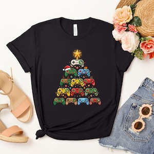May include: A black short-sleeved t-shirt featuring a graphic of a Christmas tree constructed from various colorful video game controllers. The tree is topped with a yellow star and decorated with small ornaments, a Santa hat, and reindeer antlers.