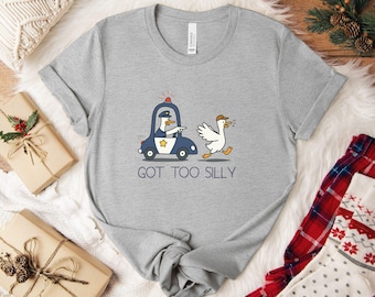 May include: A light gray t-shirt featuring a cartoon goose in a police car chasing another goose wearing a hat. The text "GOT TOO SILLY" is printed below the graphic. The shirt has a crew neck and short sleeves.