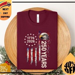 Pode incluir: Camiseta bord&ocirc; com um design patri&oacute;tico. Apresenta uma bandeira americana, uma cabe&ccedil;a de &aacute;guia e o texto "1776 2026" e "250 YEARS". A camiseta est&aacute; dobrada sobre uma superf&iacute;cie de madeira.