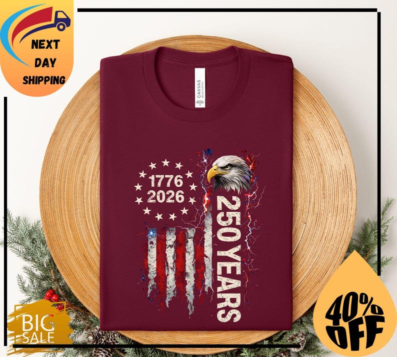 Pode incluir: Camiseta bord&ocirc; com um design patri&oacute;tico. Apresenta uma bandeira americana, uma cabe&ccedil;a de &aacute;guia e o texto "1776 2026" e "250 YEARS". A camiseta est&aacute; dobrada sobre uma superf&iacute;cie de madeira.