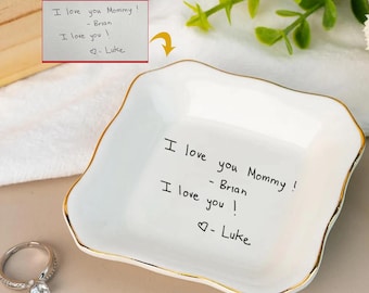 May include: A white ceramic trinket dish with a gold rim, featuring handwritten text: "I love you Mommy! - Brian, I love you! - Luke." A silver ring with a large, clear gemstone sits nearby. The dish has an irregular, wavy shape.