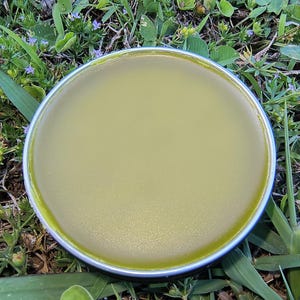 May include: A round, silver-rimmed container filled with a smooth, light green substance. The container is set on a bed of green grass and small purple flowers, creating a natural, earthy aesthetic. The product appears to be a balm or cream.