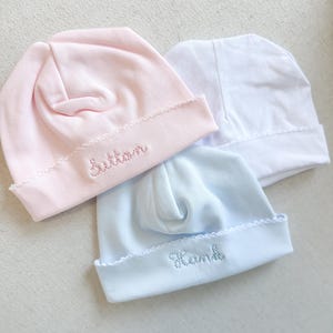 May include: Three baby hats in pink, white, and blue. The pink hat has the name "Button" embroidered on the brim, and the blue hat has "Hank" embroidered. Each hat has a scalloped edge.