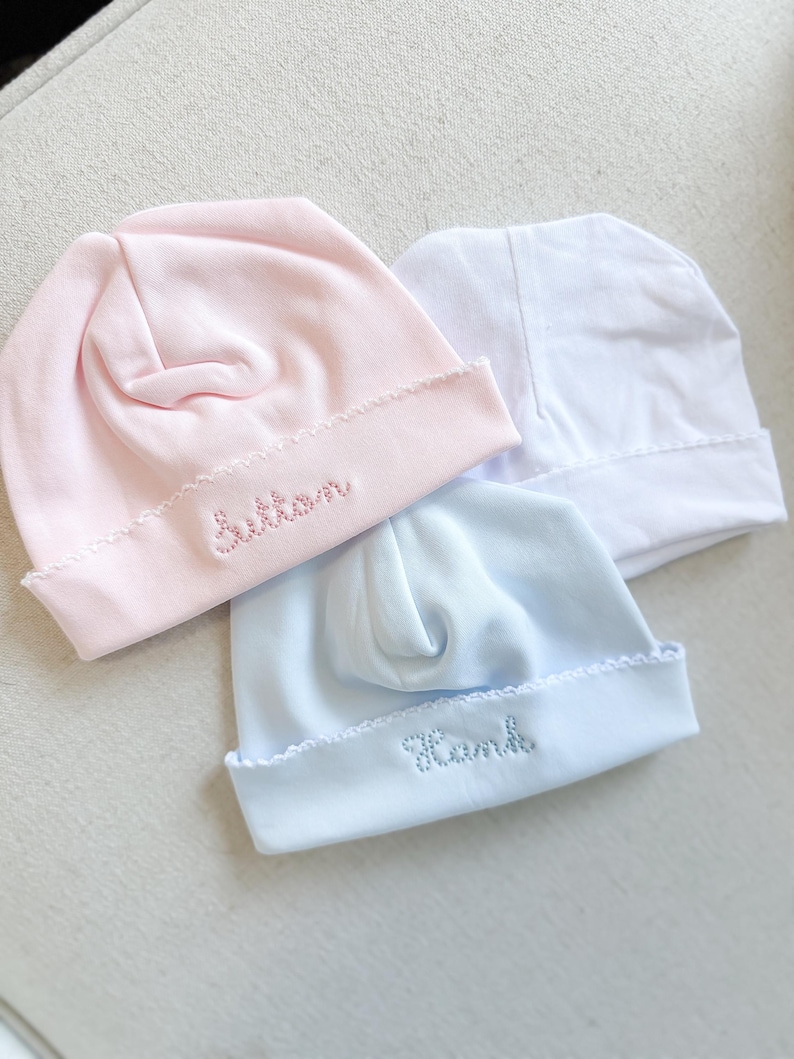 May include: Three baby hats in pink, white, and blue. The pink hat has the name "Button" embroidered on the brim, and the blue hat has "Hank" embroidered. Each hat has a scalloped edge.