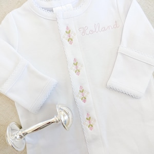 May include: A white baby onesie with the name "Holland" embroidered in pink. The onesie features pink and green floral embroidery and a silver rattle.