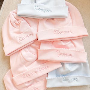 May include: A collection of baby hats in soft pastel colors, including pink and light blue. Each hat features a scalloped brim and embroidered name in a coordinating color. Names include "Cooper", "Lainey", "Eleanor", "Jenna", "Emma Grace", "Brooks", and "Bennett".