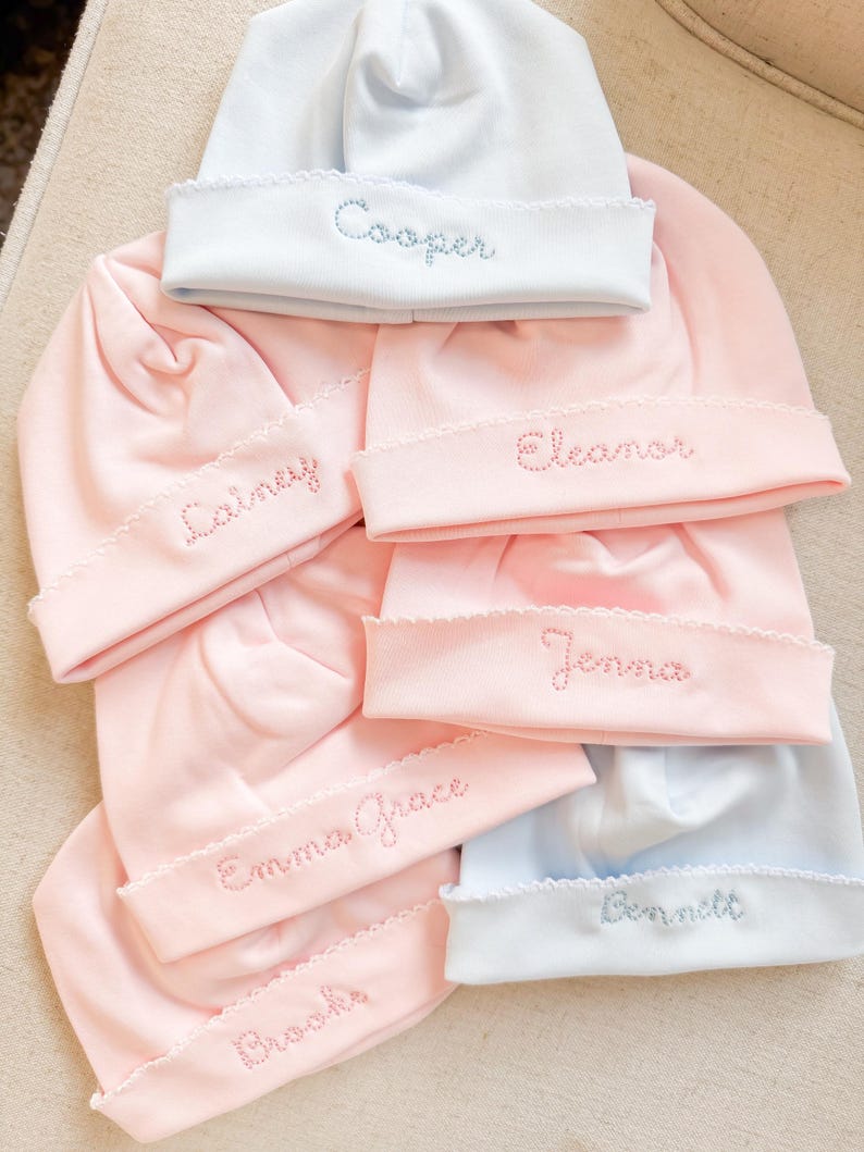 May include: A collection of baby hats in soft pastel colors, including pink and light blue. Each hat features a scalloped brim and embroidered name in a coordinating color. Names include "Cooper", "Lainey", "Eleanor", "Jenna", "Emma Grace", "Brooks", and "Bennett".