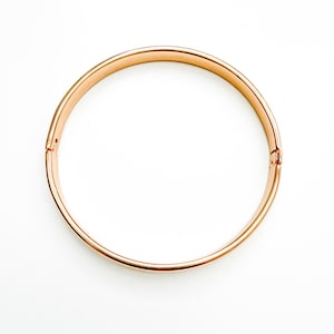 May include: A circular rose gold bangle bracelet with a smooth, polished finish. The bracelet features a hinged opening mechanism on one side and a small, secure clasp on the opposite side, creating a continuous, clean, and minimalist circular form.