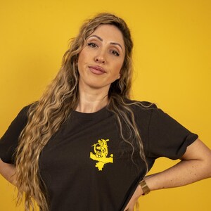May include: A woman wearing a black t-shirt with a yellow graphic design of a person holding a microphone. The woman has long, wavy blonde hair and is standing in front of a yellow background.