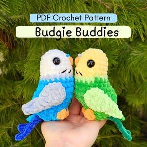 May include: Two handmade crochet parakeets, one blue and white, the other yellow and green, are shown. The birds have black eyes and orange feet. The text "PDF Crochet Pattern" and "Budgie Buddies" are displayed above the birds.