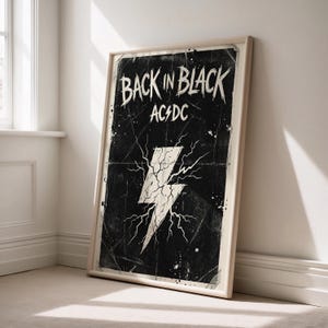 Op de afbeelding: Een ingelijste poster met een zwarte achtergrond en de tekst "BACK IN BLACK" en "AC/DC" in het wit. Een witte bliksem is gecentreerd op de verweerde zwarte achtergrond. De poster zit in een lichtgekleurde houten lijst.