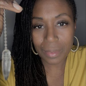 May include: A woman with long black dreadlocks wears a yellow shirt and holds a clear quartz pendulum in her hand.