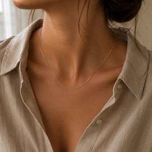 May include: A delicate gold chain necklace featuring a fine link design resting against the neck. The necklace is worn with a light beige collared shirt that has visible buttons along the front placket, creating a simple and clean aesthetic.