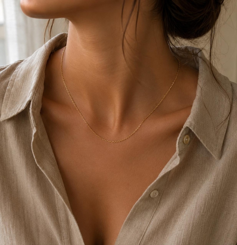 May include: A delicate gold chain necklace featuring a fine link design resting against the neck. The necklace is worn with a light beige collared shirt that has visible buttons along the front placket, creating a simple and clean aesthetic.