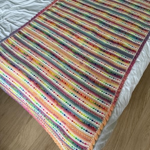 Può includere: Una coperta all'uncinetto a righe colorate con un bordo a festoni. La coperta è realizzata con una varietà di colori vivaci, tra cui rosa, arancio, giallo, verde, blu e viola.