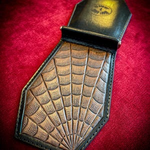 Könnte beinhalten: Eine schwarze Ledergeldbörse mit einem braunen Spinnennetz-Design. Die Geldbörse hat einen Geldclip und ist in Form eines Sarges.