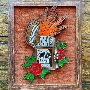 May include: A framed leather artwork depicting a vintage lighter with a skull and flames. The artwork is on a red velvet background and features green leaves and red flowers.