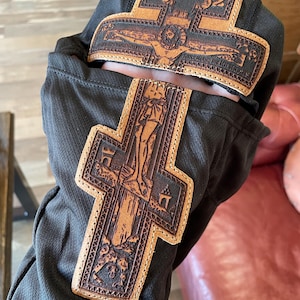 Może przedstawiać: Czarny element materiału z dwoma brązowymi sk&oacute;rzanymi krzyżami. Krzyże przedstawiają szczeg&oacute;łowe obrazy religijne i są przyszyte do materiału. Sk&oacute;ra ma jasnobrązowy kolor z ciemniejszymi brązowymi detalami.