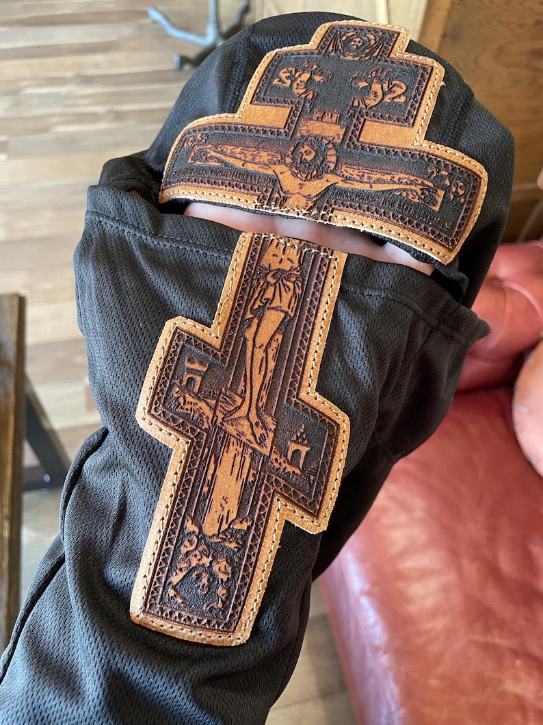 Może przedstawiać: Czarny element materiału z dwoma brązowymi sk&oacute;rzanymi krzyżami. Krzyże przedstawiają szczeg&oacute;łowe obrazy religijne i są przyszyte do materiału. Sk&oacute;ra ma jasnobrązowy kolor z ciemniejszymi brązowymi detalami.