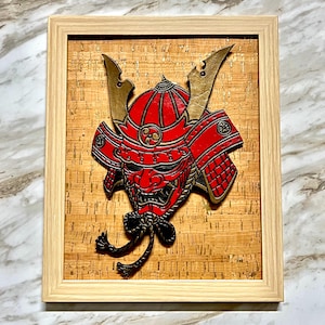 May include: Framed artwork featuring a red and gold samurai mask. The mask is intricately detailed with black accents and a decorative knot. The artwork is set against a cork background and framed in light wood.