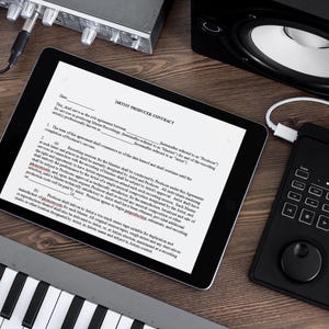May include: A black tablet with a white screen displaying a "Music Producer Contract" document. The tablet is on a wooden surface with a black speaker, a keyboard, and a black audio mixer.