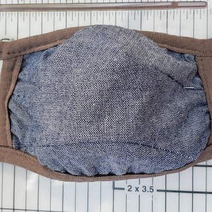 Op de afbeelding: Een bruin stoffen mondkapje met een blauwe denim voering. Het masker heeft twee bruine stoffen bandjes.