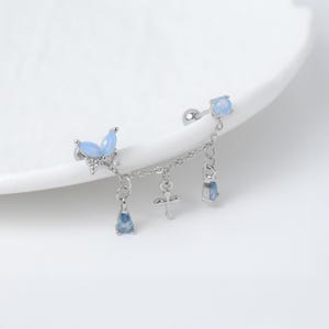 May include: A silver-toned cartilage earring featuring a butterfly stud with light blue stones, connected by a delicate chain to a second stud. Three charms hang from the chain: two light blue teardrop-shaped stones and a small silver cross.