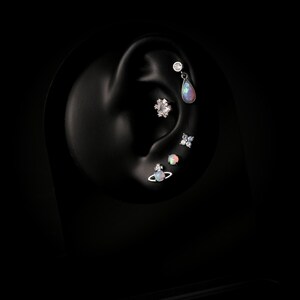 May include: A set of five silver-toned ear piercings displayed on a black ear model. The jewelry includes a teardrop opal dangle, a floral crystal cluster, a four-stone crystal flower, a round opal stud, and a Saturn-shaped opal planet stud.