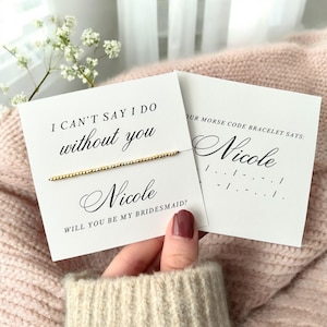 May include: A white card with the text "I can't say I do without you Nicole Will you be my bridesmaid?" and a gold beaded bracelet. A second white card says "Our Morse code bracelet says: Nicole" with the Morse code for the name Nicole.