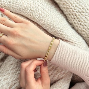 May include: A gold beaded bracelet with two strands of small, round beads. The bracelet is worn on a person's wrist.