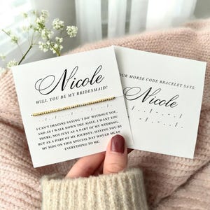 May include: A white card with black text asking "Will you be my bridesmaid?" with the name "Nicole" written in cursive. A gold bracelet with a message in Morse code is attached to the card. The card also includes a message about the importance of friendship and support.