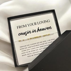 May include: A black gift box with a white card that reads "FROM YOUR LOVING cousin in heaven." A delicate beaded bracelet is displayed on the card. The bracelet has gold and white beads. The box is open, revealing the contents.
