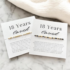 May include: Two white cards with black text that reads "18 Years Loved" and a message about being loved through every joy and challenge. Each card has a bracelet made of small white, black, and gold beads.