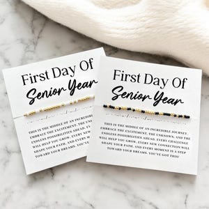 Peut inclure: Deux cartes blanches avec le texte "First Day Of Senior Year" et un bracelet de perles. Un bracelet a des perles dorées et blanches, l'autre a des perles noires et dorées. Les cartes ont un texte inspirant.