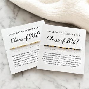 May include: Two white cards with the text "First Day of Senior Year Class of 2027" and a beaded bracelet. One bracelet has white and gold beads, the other has black and gold beads. The cards have inspirational text.