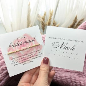 May include: A white card with a pink watercolor rose design and the text "To my bridesmaid" and a message about how grateful the person is to have their bridesmaid. The card also includes a gold bracelet with a message in Morse code. The card says "Your Morse Code Bracelet Says: Nicole" and shows the Morse code for the name Nicole.