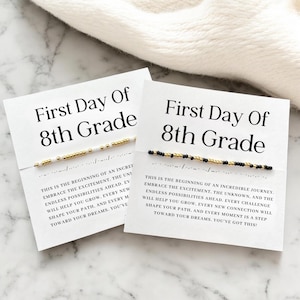 May include: Two white cards with the text "First Day Of 8th Grade" and a beaded bracelet. The cards also have inspirational text. One bracelet has gold and white beads, the other has black and gold beads.